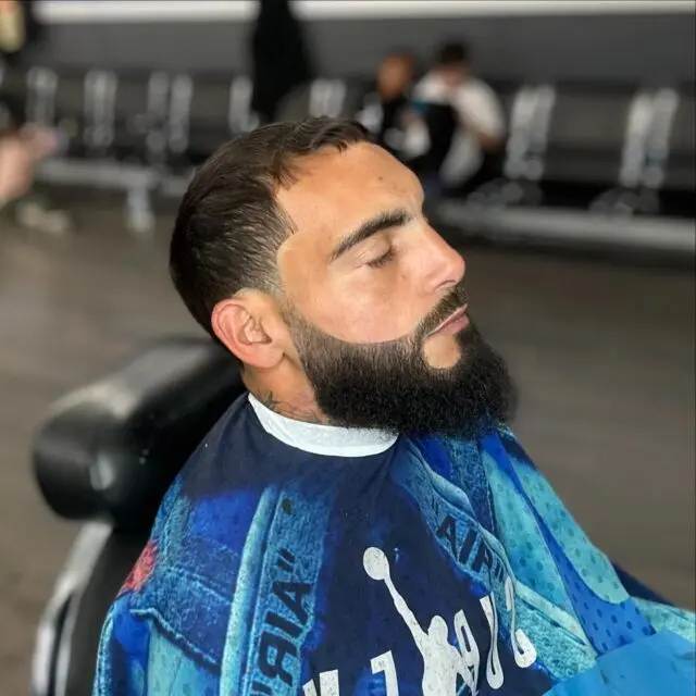 A client with a sculpted full beard and fade relaxing in the chair